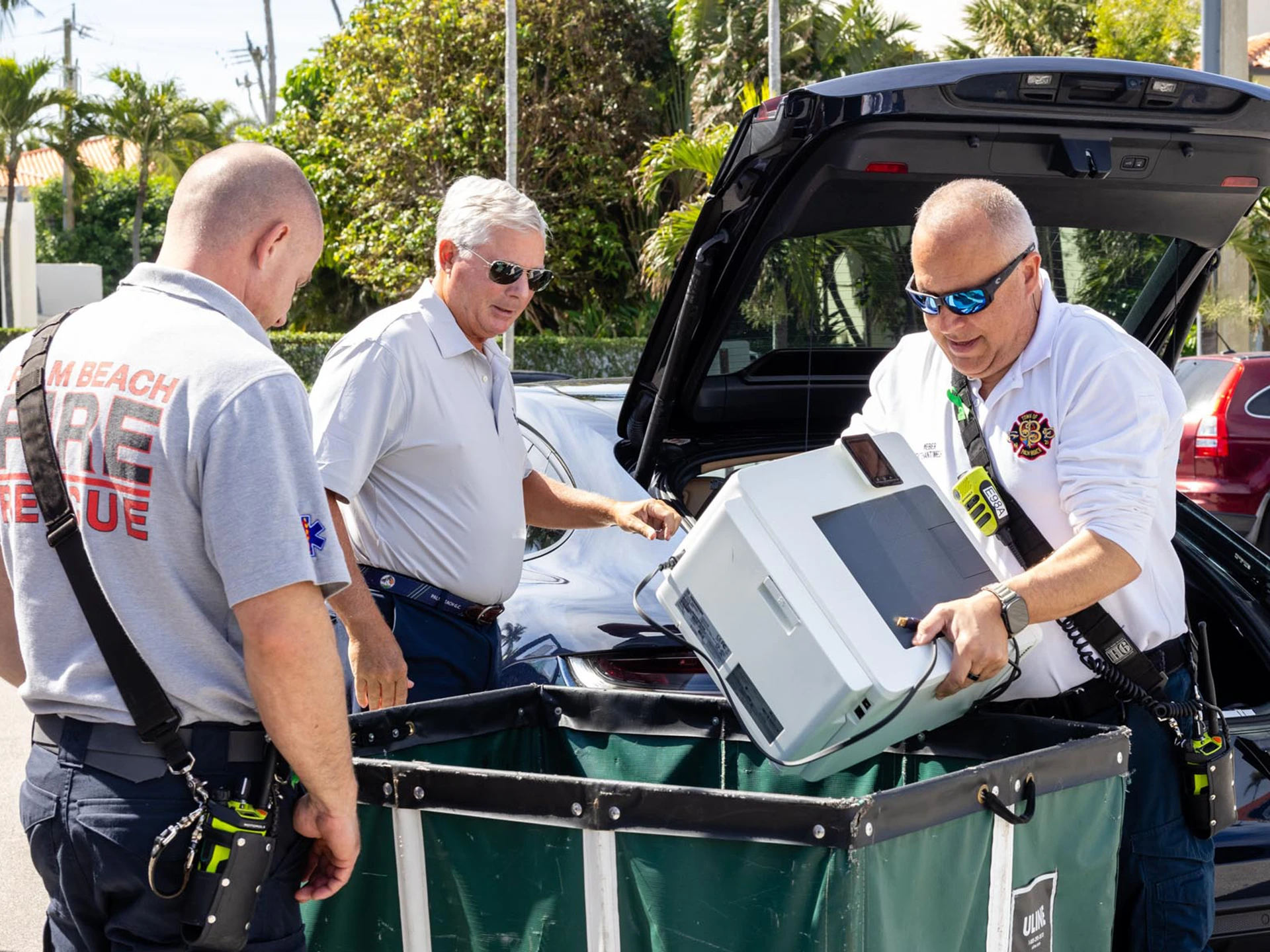 Three men in white shirts and sunglasses load a printer into a green bin at the Palm Beach Police & Fire Foundation Safeguard Community Cleanout Event.